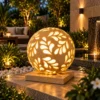 Handcrafted sandstone garden light ball with carved floral pattern glowing warmly on a stone base outdoors